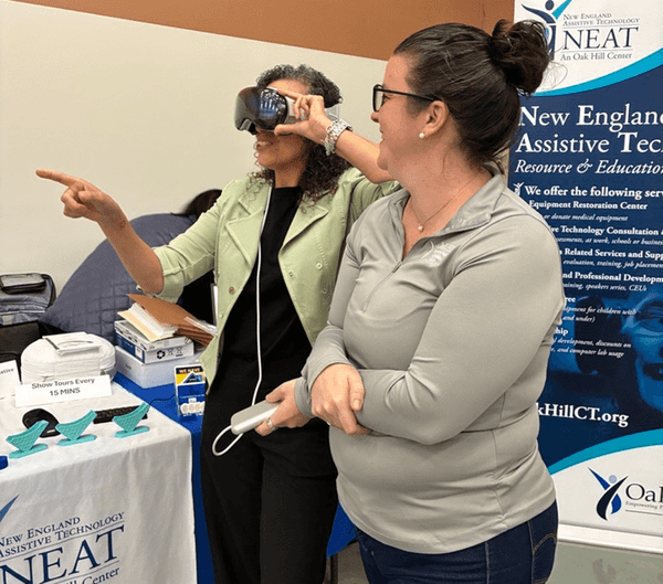 Two women at a booth; one is wearing a headset and the other is assisting her while standing next to a table with informational materials and a NEAT Center banner.