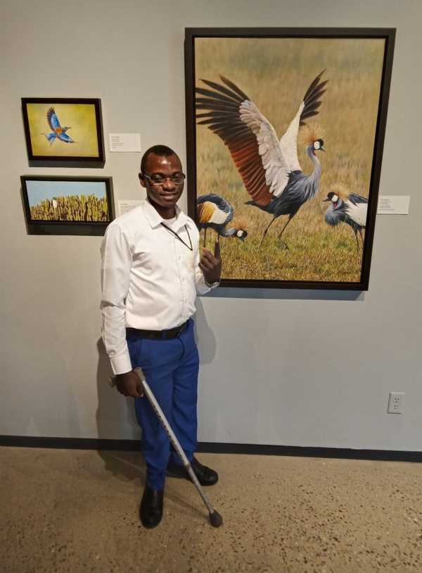 Harunah wearing a white shirt and blue pants standing near a painting of a bird pointing to it with his thumb and holding a crutch in his other hand.